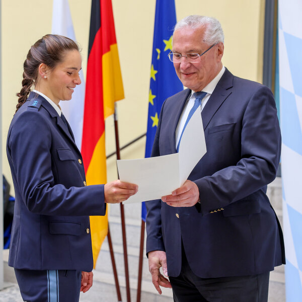 Innenminister Joachim Herrmann überreicht einer Polizeisportathletin ihre Auszeichnungsurkunde vor einem Fahnenset im Odeon des Bayerischen Innenminiserium