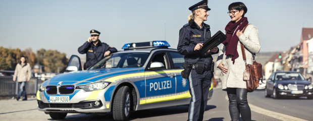 Eine Polizistin unterhält sich mit einer Frau. Dahinter ist ein Polizei-Auto zu sehen. Und ein Polizist.