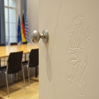 Geöffnete Tür Das Foto zeigt einen Ausschnitt eines Sitzungssraums, im Hintergrund sind die Fahnen Bayerns, Deutschlands und die Europaflagge ersichtlich.