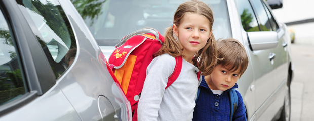 Zwei Schulkinder stehen zwischen geparkten Autos.